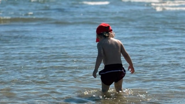 Ein kleines blondes Kind, von hinten gesehen, mit einer roten Mütze, geht an einem sonnigen Tag allein ins Meer, das Wasser reicht ihm bis zu den Waden.