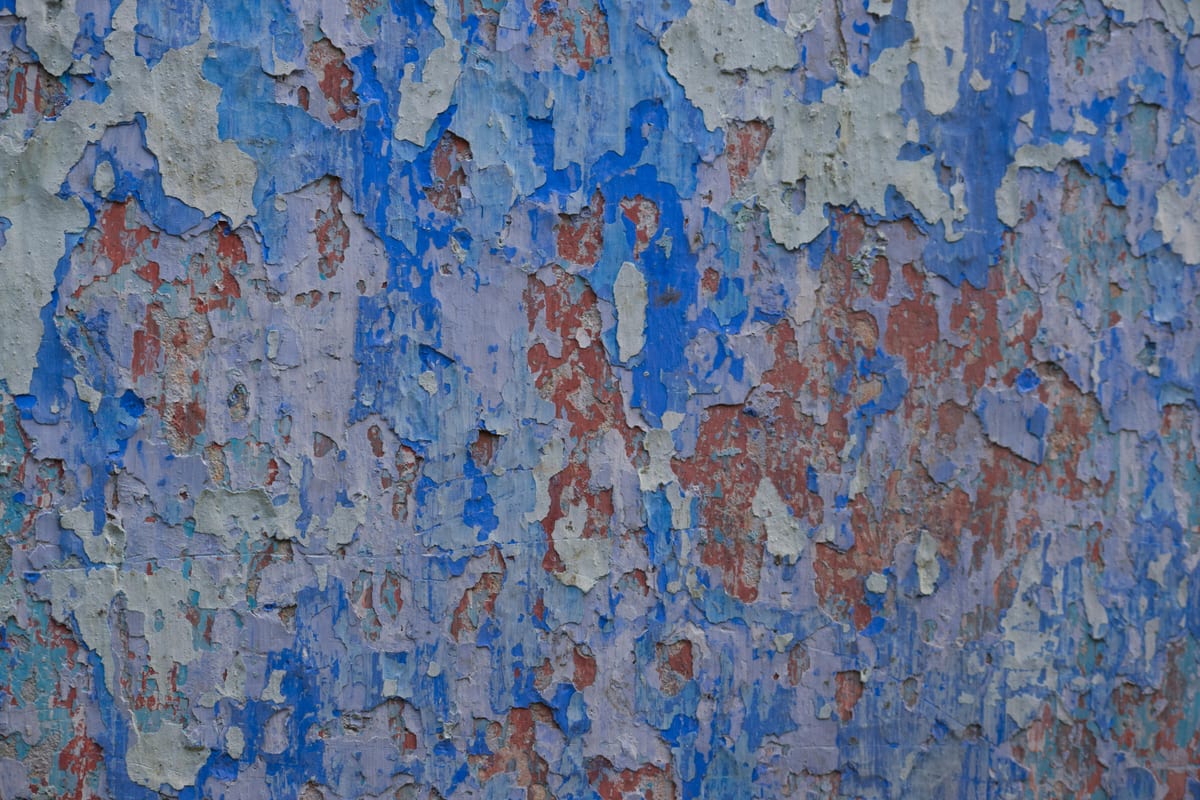 Eine Wand mit abplatzender Farbe in mehreren Schichten — Blau, Grau, Rost — über die Jahre übereinandergelegt.