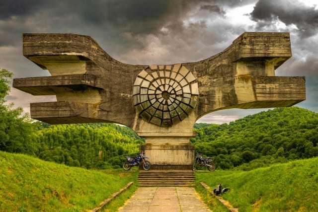 Un monumento brutalista di cemento si erge contro un cielo carico di nuvole, due motociclette parcheggiate sotto l'arco. Un disco scolpito al centro come un orologio fermo. Il documento che descrive un sistema che non c'è più.