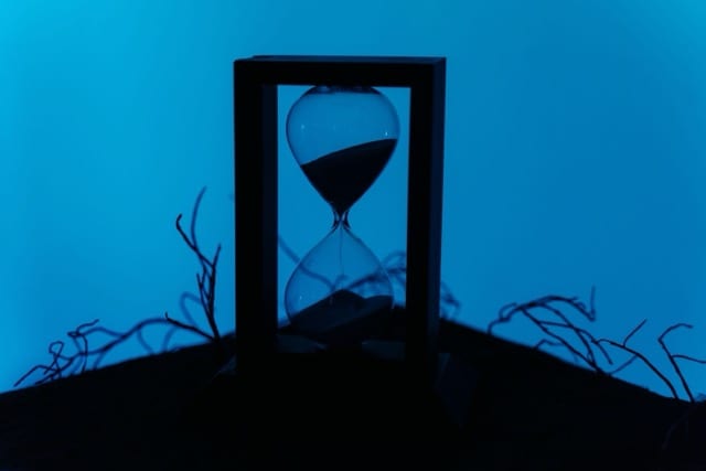 A glass-and-metal hourglass backlit against a midnight-blue background, resting on a dark surface among dry, twisting branches. The pale sand is sliding toward the lower bulb. Time is passing, but it keeps flowing: it hasn't stopped.