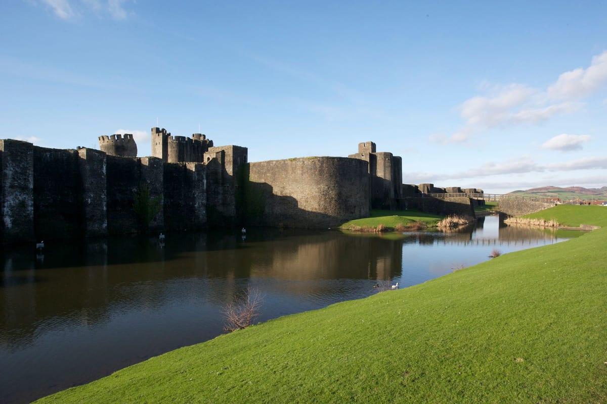 Caerphilly Castle in Wales, gesehen von jenseits seines Burggrabens: eine grüne Wiese im Vordergrund, stilles Wasser dazwischen, graue Mauern am anderen Ufer. Der Graben ist der größte Teil der Befestigung, und zugleich der Teil, der nie gekämpft hat.