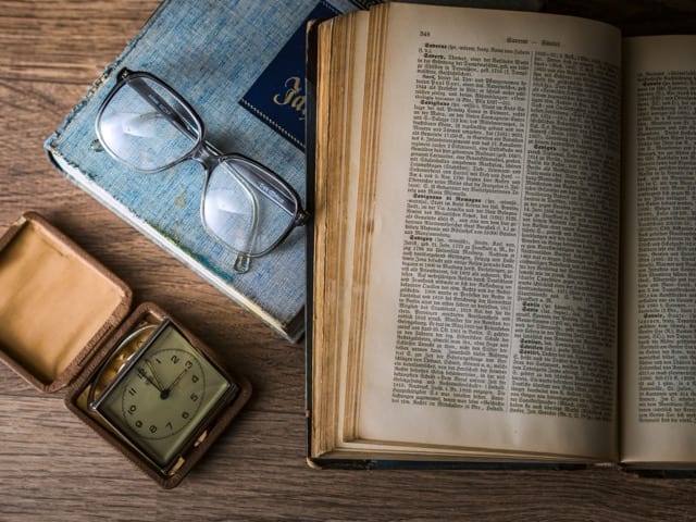 An old dictionary open on a page dense with entries, resting on a wooden table. Beside it, a pair of vintage glasses laid on a worn book with a blue cover and a small travel clock in its leather case. Words, reading, and time, the tools with which a language decides its own future.