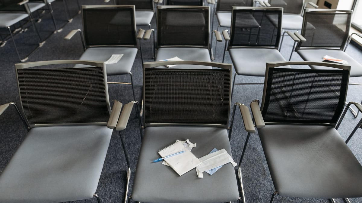 Reihen identischer Stühle in einem leeren Konferenzraum, von oben gesehen. Auf einem einzigen Stuhl in der Mitte liegen ein aufgeschlagener Notizblock, ein Stift und ein paar Blätter: der einzige Platz, an dem wirklich jemand war.