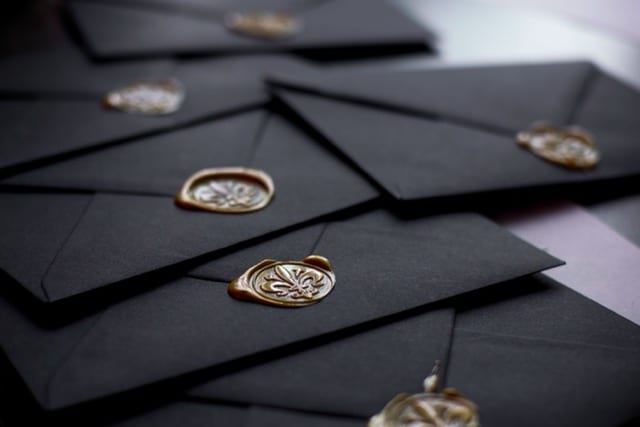 Ein Stapel schwarzer Umschläge, fächerförmig auf einer dunklen Fläche angeordnet, jeder mit einem goldenen Wachssiegel verschlossen, in das ein Wappen geprägt ist. Der Fokus liegt auf einem Siegel im Vordergrund, die übrigen verschwimmen im Hintergrund. Die alte Geste, die Herkunft dessen zu verbürgen, was darin verschlossen ist.