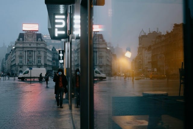 Una strada europea al crepuscolo, vista da una pensilina di vetro. Una persona cammina nella luce arancione delle insegne accese e dei lampioni. La sua immagine si riflette nel vetro accanto, sdoppiata. Lo spettro è il riflesso, è chi guarda, è entrambi.