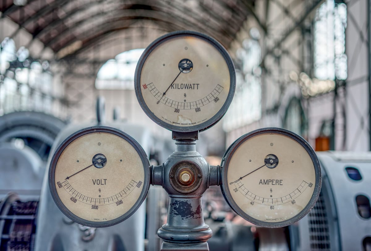 Drei historische Messinstrumente in einer industriellen Maschinenhalle: ein Voltmeter, ein Amperemeter und ein Kilowattzähler auf einer grauen Metallsäule, die Zeiger in Ruhestellung. Die Instrumente sind perfekt lesbar; der Strom, den sie messen sollten, fließt nicht mehr.