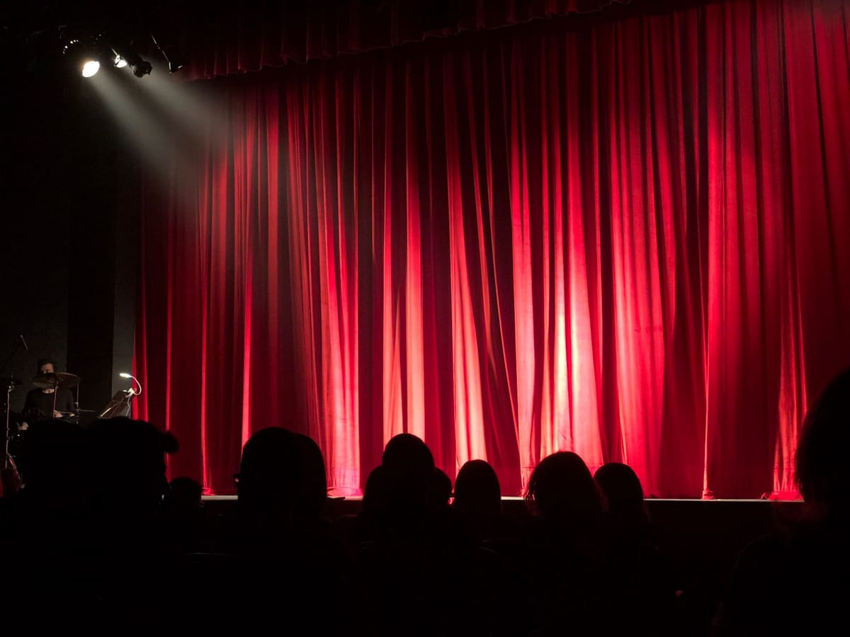 Geschlossener roter Bühnenvorhang in einem Theater, beleuchtet von einem einzelnen Scheinwerfer, davor die Silhouetten eines bereits sitzenden Publikums. Keine Generalprobe: Wenn der Vorhang aufgeht, ist die erste Vorstellung die Premiere.