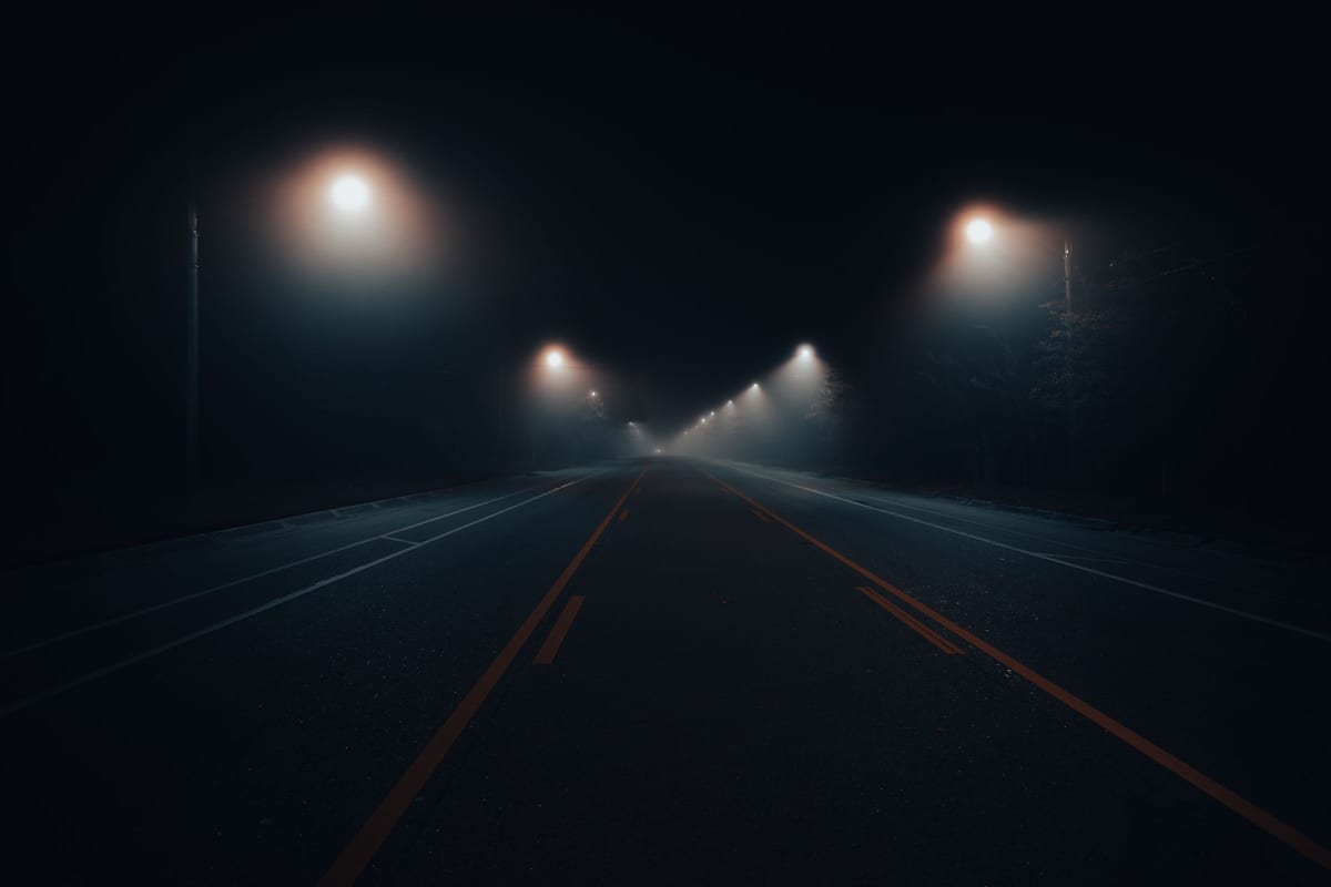 A completely empty two-lane road at night, in fog. Yellow and white lane markings converge toward a dark vanishing point under a few streetlights fading into the distance. No cars, no people.