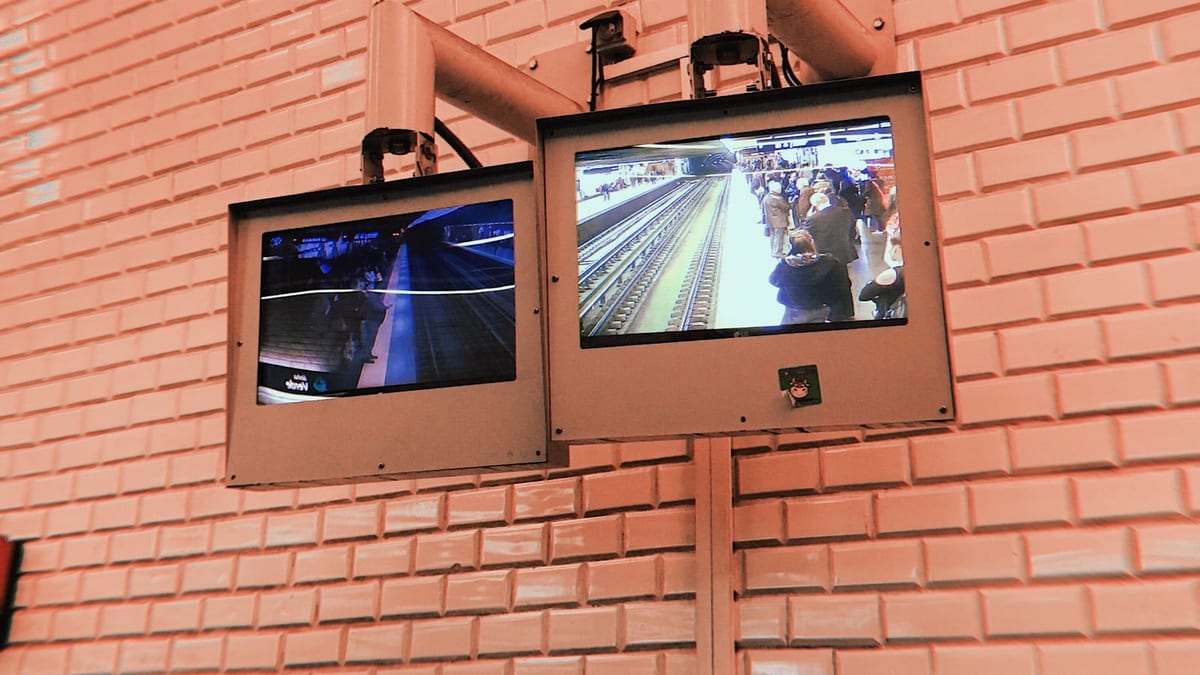 Zwei Überwachungsmonitore, hoch an einer rosafarbenen Kachelwand in einer U-Bahn-Station montiert. Der eine zeigt einen von Reisenden überfüllten Bahnsteig, der andere einen leeren Bahnsteig im Halbdunkel. Über den Monitoren sind die Kameras befestigt. Niemand sieht auf die Bildschirme.