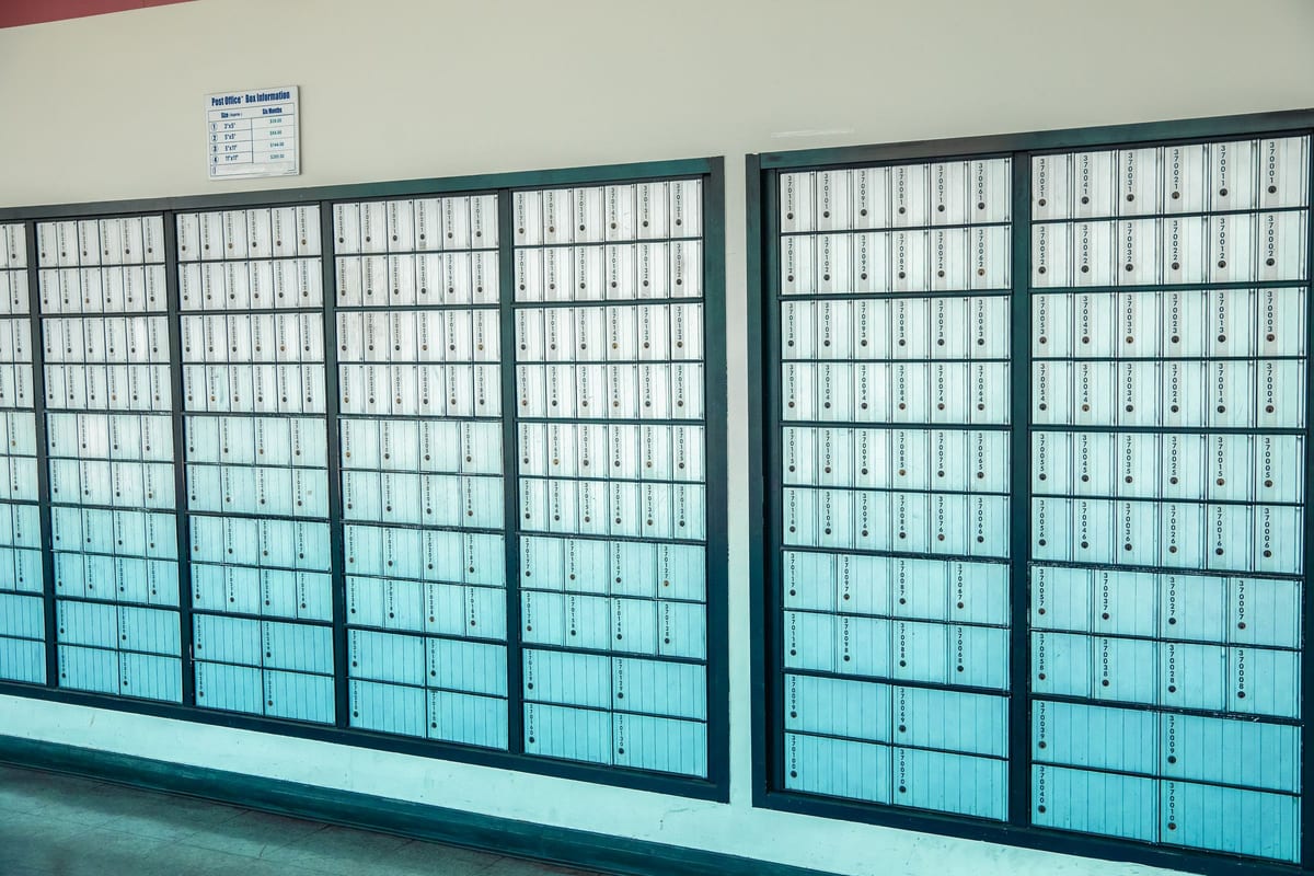 A post office wall filled end to end with hundreds of identical white and teal metal boxes, gridded behind dark frames, each with its own lock and number. None of them is worth much on its own.